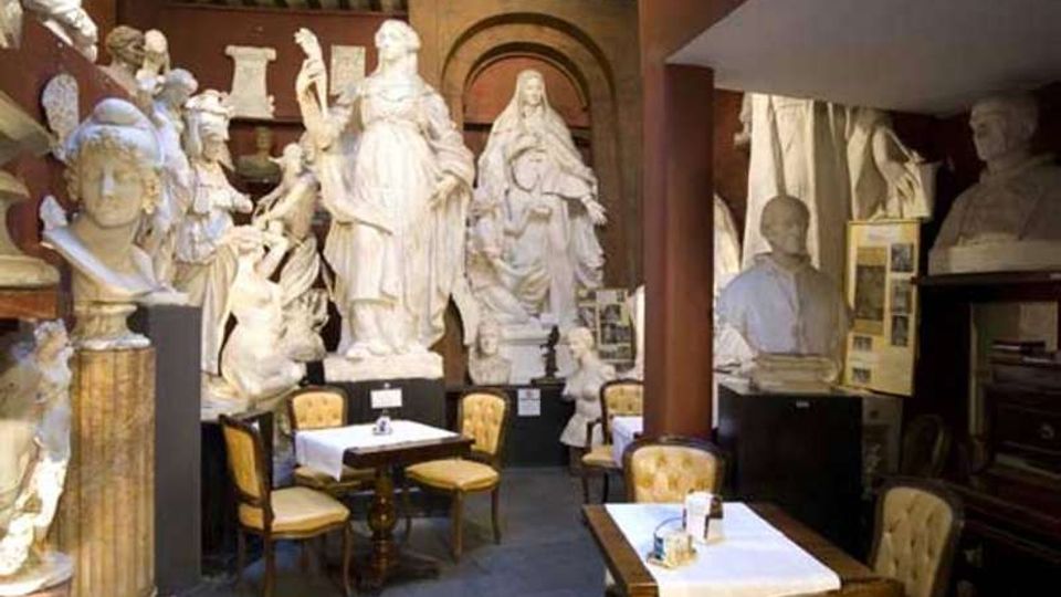 Interno di un locale simile a un caffè-museo con tavolini apparecchiati e sedie imbottite, circondato da numerose grandi statue e busti in gesso disposti lungo le pareti. Soffitto con travi a vista, colonne in legno e pareti ricoperte di sculture e pannelli informativi.