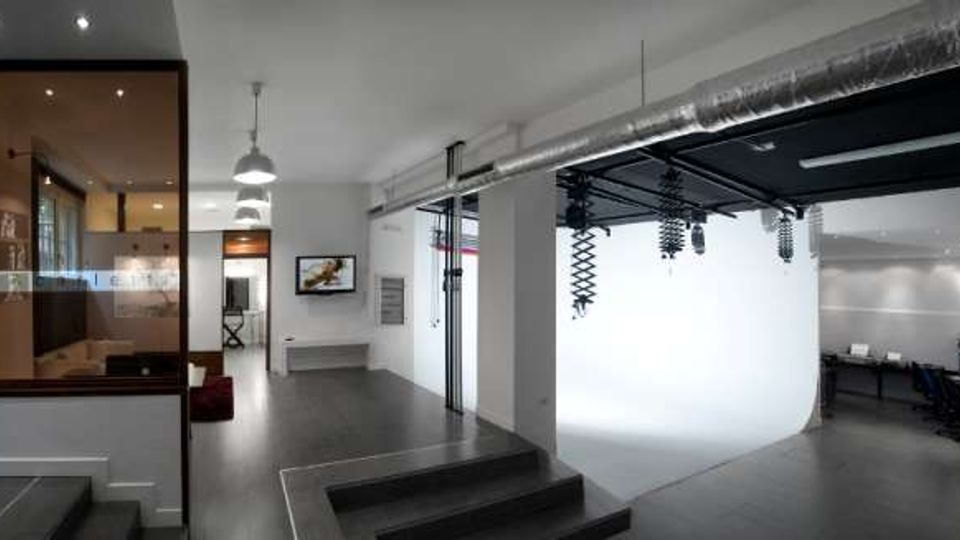 Interno di uno studio fotografico moderno con pavimento scuro, pareti bianche e condotte a vista sul soffitto. A destra si vede un’area set con fondale bianco curvo e luci sospese, mentre a sinistra c’è una sala separata da una parete vetrata e sul fondo una zona lavoro con tavoli e sedie.