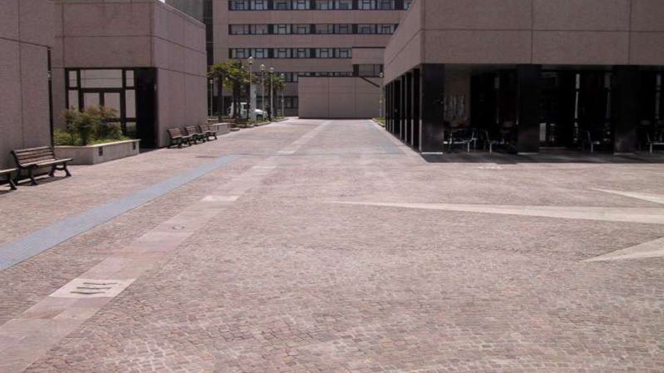 Ampio piazzale pavimentato in pietra tra edifici moderni in cemento, con panchine lungo il lato sinistro e lampioni. A destra un portico con colonne crea una zona coperta, mentre sullo sfondo si vede un edificio a più piani con finestre orizzontali.