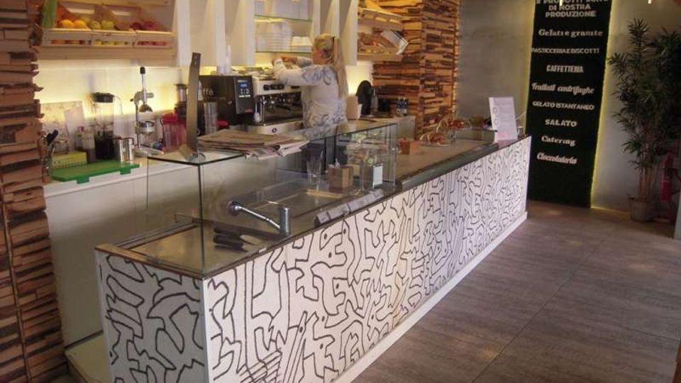 Interno di una caffetteria/gelateria moderna con lungo bancone vetrato e frontale bianco decorato a linee, pareti con rivestimento in listelli di legno e scaffali illuminati con prodotti. Sullo sfondo si vede l’area macchina del caffè e una parete scura con scritte del menù.
