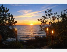 Vista sul mare al tramonto da un giardino costiero, con vegetazione in primo piano e torce accese su pali lungo il bordo. All’orizzonte il sole scende sull’acqua calma sotto un cielo con nuvole sottili.
