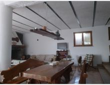 Soggiorno rustico con grande tavolo in legno e panche, soffitto alto con travi scure a vista e mensola sospesa. Sullo sfondo si vedono un camino ad angolo, una zona divani e sedie, una TV su mobile e una finestra rettangolare con cornice in legno.