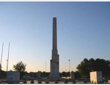 Obelisco in pietra su alto basamento al centro di una rotonda o piazza, circondato da piccoli arbusti e blocchi squadrati. Sullo sfondo si vedono alberi, alcuni lampioni stradali e un cielo sereno al tramonto.