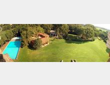 Vista aerea di una tenuta con grande prato verde circondato da alberi, edifici con tetti in terracotta e una piscina rettangolare azzurra sul lato sinistro. Sullo sfondo si vedono aree boscate e campi aperti.