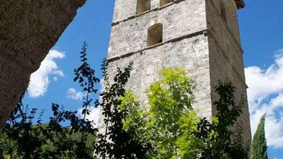 Torre campanaria in pietra con tetto a falde e aperture ad arco, vista dal basso tra alberi e vegetazione. Sullo sfondo si vede il cielo azzurro con alcune nuvole e una collina boscosa.