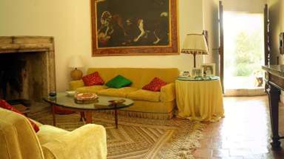 Soggiorno luminoso con divani chiari e cuscini colorati, camino in pietra, tavolino rotondo e lampade da tavolo. Pavimento in parquet e grande apertura vetrata che conduce all’esterno, con una consolle in legno lungo la parete.