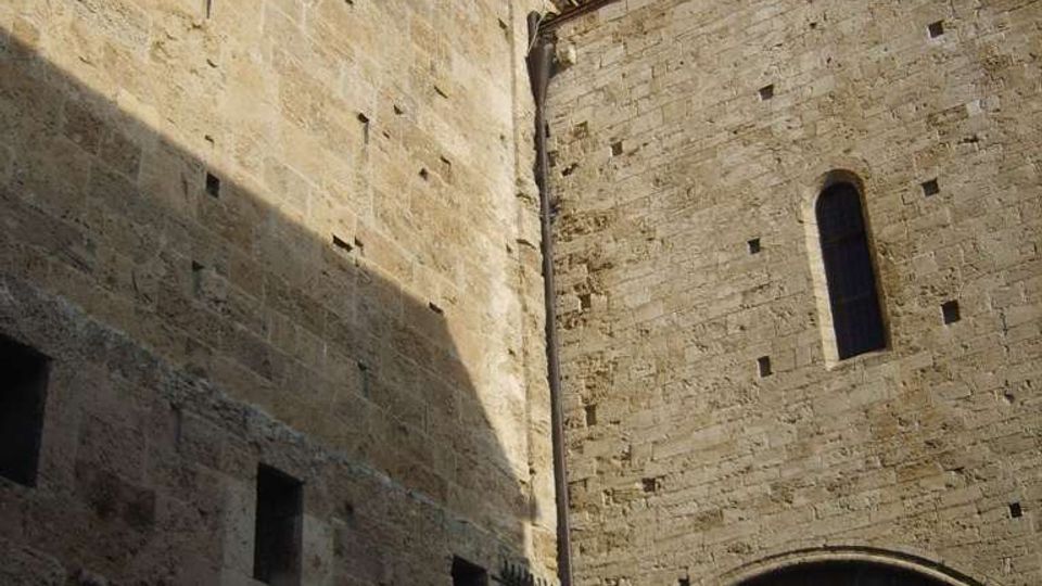 Facciata in pietra di un edificio storico, con rosone circolare decorato e piccole aperture rettangolari. A destra si vede un grande arco d’ingresso con cancello in ferro e una finestra stretta ad arco sotto un tetto in tegole, sullo sfondo cielo parzialmente nuvoloso.