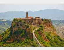 Borgo storico in pietra arroccato su una rupe di tufo, con campanile e case compatte attorno a un ingresso ad arco. Un lungo ponte pedonale sopraelevato collega il paese al crinale, con colline verdi e montagne sullo sfondo.