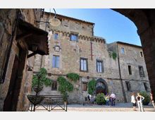 Facciata di un palazzo storico in pietra affacciato su una piccola piazza lastricata, con finestre rettangolari e portale ad arco al centro. La scena è inquadrata da un arco in primo piano e include una lampada a muro e vegetazione rampicante sulle pareti.