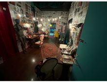 Interno di un camerino teatrale con pareti e soffitto rivestiti di pagine di giornale e una fila di lampadine lungo i lati. Specchi con cornici dorate, tavoli da trucco pieni di oggetti, manichini, sedie e un tappeto centrale conducono a una porta in fondo che si apre su un corridoio illuminato.