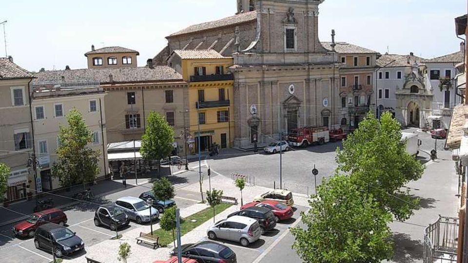 Piazza cittadina con parcheggi, aiuole e alberi, circondata da edifici a più piani con tetti in coppi. Sullo sfondo domina una grande chiesa in pietra con facciata classica, campanile e portale centrale.