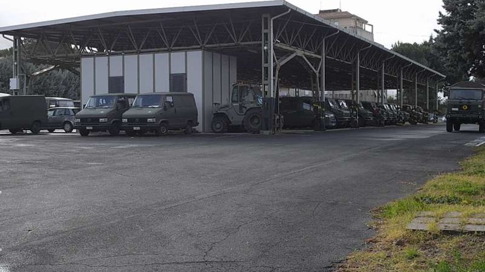 Ampia tettoia industriale in metallo con struttura a travi e pilastri, affiancata da un edificio prefabbricato a pannelli. Sotto la copertura sono parcheggiati diversi veicoli e mezzi, con grande piazzale asfaltato e alberi sullo sfondo.