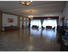 Ampia sala interna con pavimento in piastrelle e lampadari a soffitto, arredata con divani e poltrone disposti lungo le pareti. Grandi finestre con tende e drappeggi occupano il fondo della stanza, con elementi decorativi e piante ai lati.