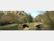 Ponte in pietra a due arcate che attraversa un fiume calmo, circondato da alberi spogli e vegetazione lungo le rive. Sullo sfondo si vedono case in pietra su un pendio e colline boscose.
