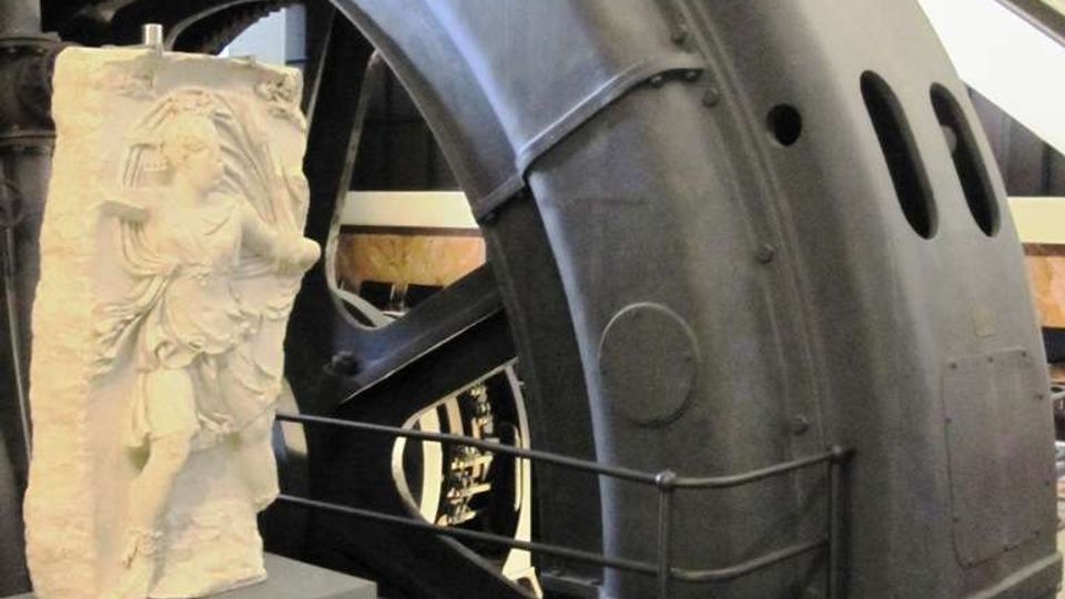 Interno di un museo tecnico con una grande turbina o generatore industriale in metallo scuro, con carter curvo forato e passerelle con ringhiere sullo sfondo. In primo piano c’è un piedistallo con un bassorilievo in pietra esposto vicino al macchinario.