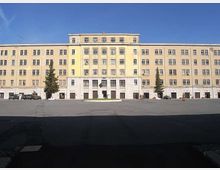 Ampia piazza asfaltata davanti a un grande edificio istituzionale rettangolare a più piani, con facciata color crema, molte finestre regolari e ingresso centrale con colonne. Ai lati dell’ingresso si vedono alcuni alberi alti e il cielo sereno sullo sfondo.