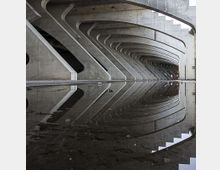 Sottopasso in cemento con una serie di pilastri e archi curvi ripetuti lungo il soffitto, in prospettiva. Una grande pozzanghera in primo piano riflette la struttura e le linee dell’architettura.
