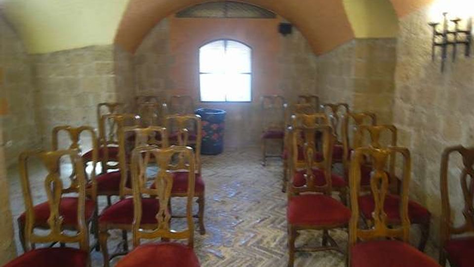 Piccola sala interna con soffitto a volta e pareti in pietra, arredata con file di sedie in legno con seduta rossa su pavimento in mattoni a spina di pesce. In fondo c’è una finestra ad arco che illumina l’ambiente e una lampada a parete in ferro.