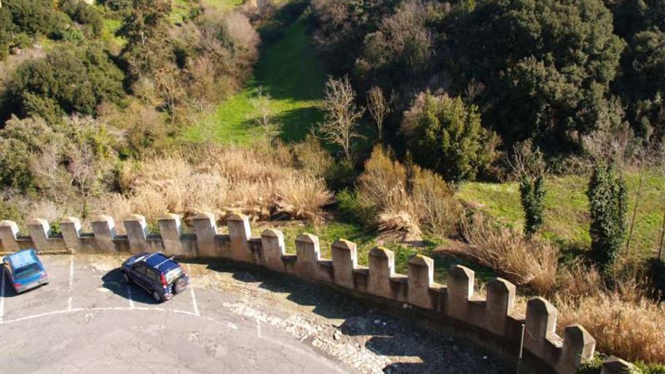 Parcheggio asfaltato curvo affacciato su una valle verde, delimitato da un muretto in pietra con merlature. Sullo sfondo si vede un pendio boscoso con alberi e cespugli e un’area di prato che si apre tra la vegetazione.