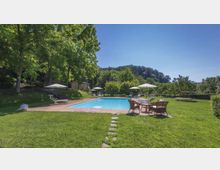 Piscina all’aperto rettangolare circondata da un ampio prato verde con sedie a sdraio, tavoli e ombrelloni. Sullo sfondo si vedono alberi alti, una collina boscosa e alcune piccole strutture in muratura ai margini del giardino.