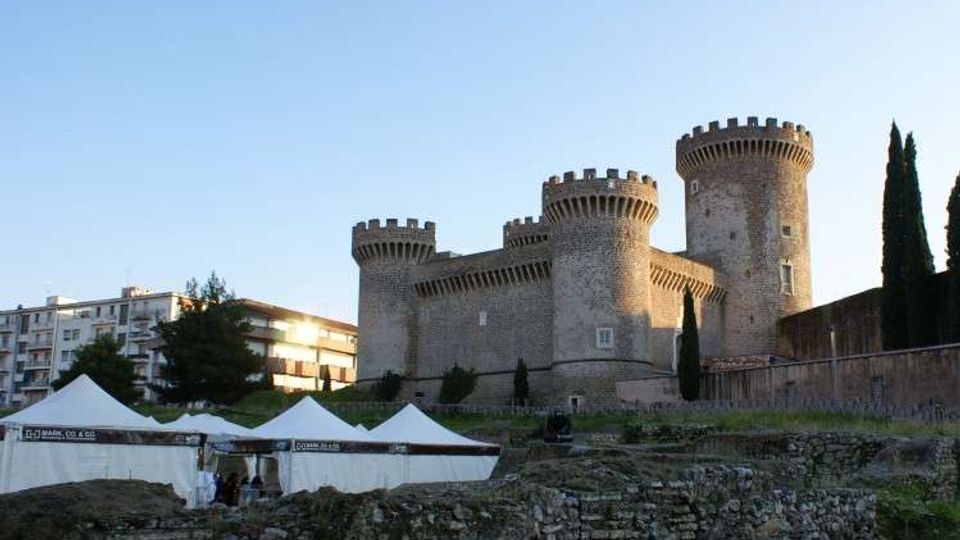 Castello in pietra con torri cilindriche merlate su un lieve rialzo, affiancato da cipressi e mura. In primo piano si vedono rovine in muratura e alcune tensostrutture bianche, con palazzi moderni sullo sfondo.