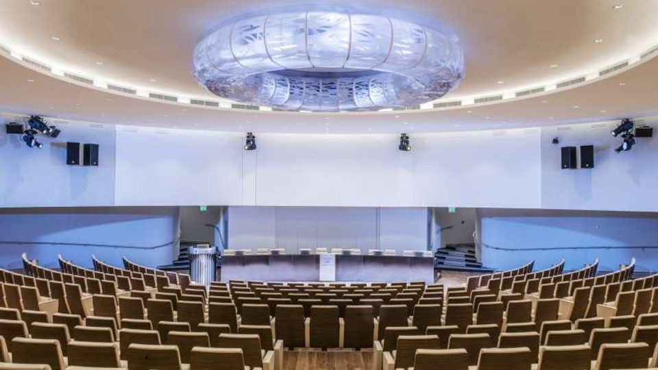 Auditorium moderno con file di poltrone in legno e tessuto disposte a gradoni, rivolte verso un palco con grande schermo bianco e podio centrale. Soffitto curvo con ampia struttura circolare luminosa e impianti audio e luci laterali.