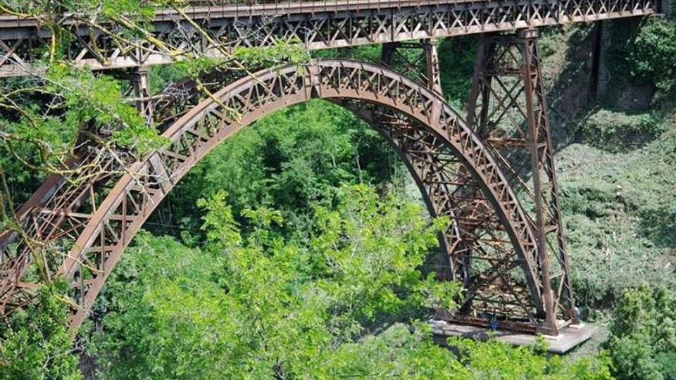 Ponte ad arco in ferro con struttura a traliccio che attraversa una gola, sostenuto da piloni metallici. Intorno si vede una fitta vegetazione e il pendio roccioso della valle.