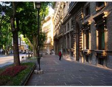 Marciapiede largo in una strada urbana alberata, con aiuole fiorite, lampioni e segnaletica stradale lungo il bordo. Sul lato destro si affacciano edifici storici in muratura con finestre alte e cornici decorative, illuminati dalla luce laterale del sole.