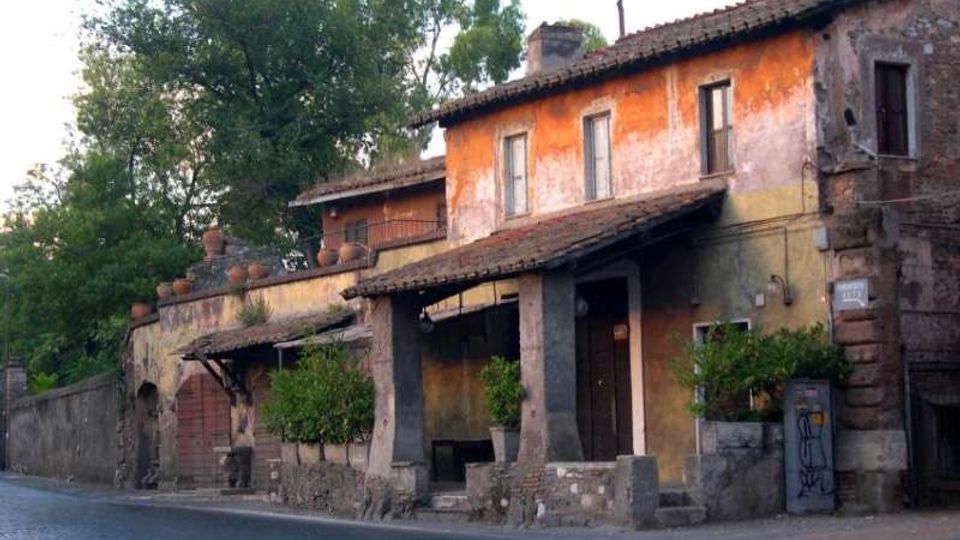 Casa rurale in pietra e intonaco color ocra, con tetto in tegole e piccolo portico su pilastri, affacciata su una strada asfaltata. Sul lato si vedono un cancello, vasi e muretti bassi, con alberi alti sullo sfondo.
