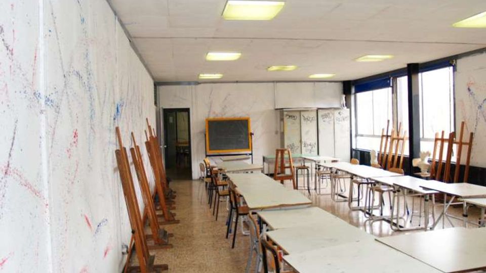 Aula scolastica adibita a laboratorio d’arte con lunghi tavoli e sedie in fila, cavalletti di legno appoggiati alle pareti e una lavagna in fondo. Grandi finestre su un lato illuminano l’ambiente, con pareti segnate da schizzi e un soffitto con pannelli luminosi.
