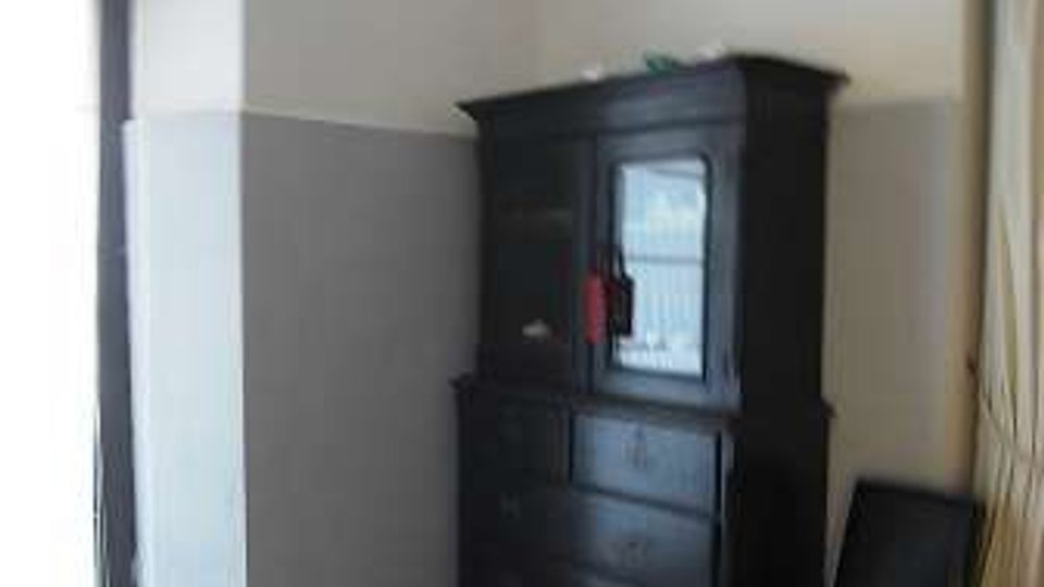 Interno di una stanza spoglia con pareti bianche e grigie e pavimento in graniglia, illuminata da una porta finestra sulla sinistra. Nell’angolo c’è una credenza scura in legno e, accanto, una sedia nera; dal soffitto pende una lampada a sospensione.