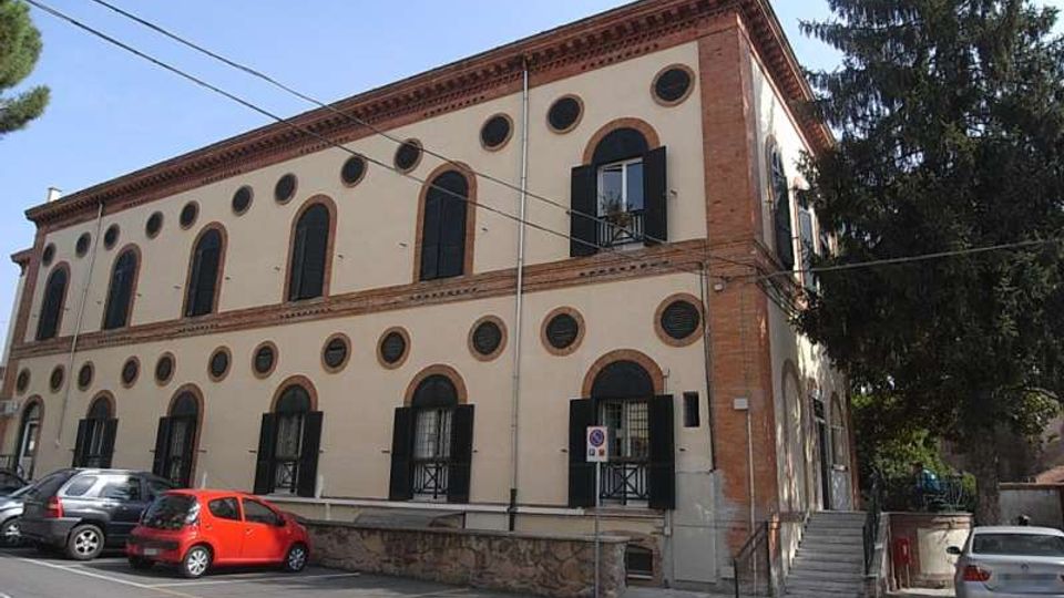 Edificio a due piani in stile storico con facciata intonacata chiara, bordi in mattoni e numerose finestre ad arco e oculi circolari con scuri scuri. Davanti c’è un parcheggio su strada e, sul lato destro, una scalinata esterna accanto ad alberi alti.