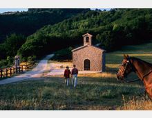 Piccola chiesa rurale in pietra con campanile a vela e portale ad arco, situata in un prato lungo una strada sterrata con staccionate. Sullo sfondo, colline ricoperte di bosco e campi aperti.