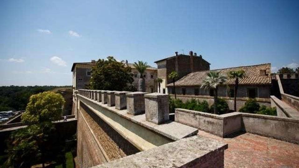 Terrazza panoramica in pietra su bastioni murati, con parapetto scandito da pilastri e pavimentazione in mattoni. Sullo sfondo si vedono edifici storici con tetti in coppi, palme e alberi, sotto un cielo sereno.