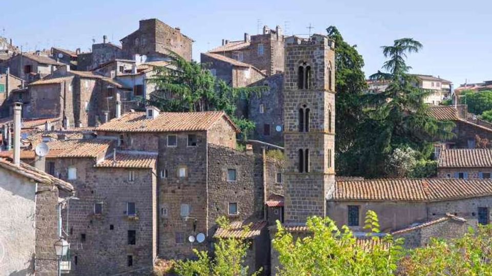 Veduta di un borgo storico con case in pietra e tetti in coppi, addossate su un pendio. Al centro spicca una torre campanaria in pietra con finestre ad arco, circondata da alberi e vegetazione.