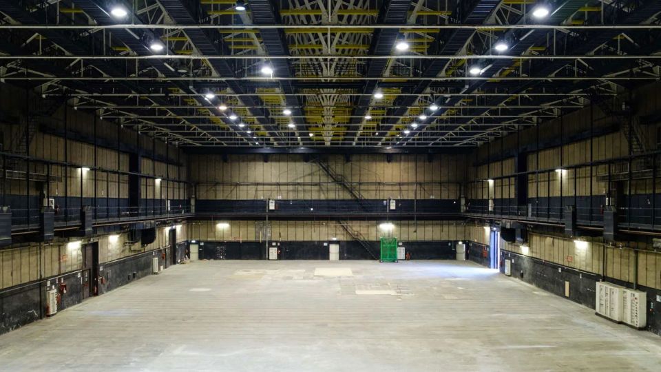Grande capannone industriale vuoto con ampia pavimentazione in cemento e pareti scure, illuminato da numerosi faretti a soffitto. Struttura metallica a traliccio con passerelle laterali sopraelevate e porte di servizio lungo i lati.