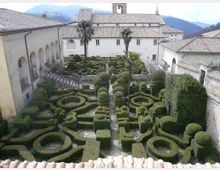 Veduta dall’alto di un chiostro con giardino formale all’italiana, caratterizzato da siepi potate in forme geometriche e vialetti centrali. Il cortile è circondato da edifici in muratura chiara con tetti in coppi e un campanile sullo sfondo, con montagne in lontananza.