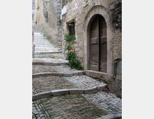 Vicolo in pietra acciottolata in un borgo storico, con pavimentazione a gradoni che sale lungo muri in muratura grezza. A destra si vede un grande portale ad arco con doppia porta in legno, affiancato da piccole piante e una finestra incassata.