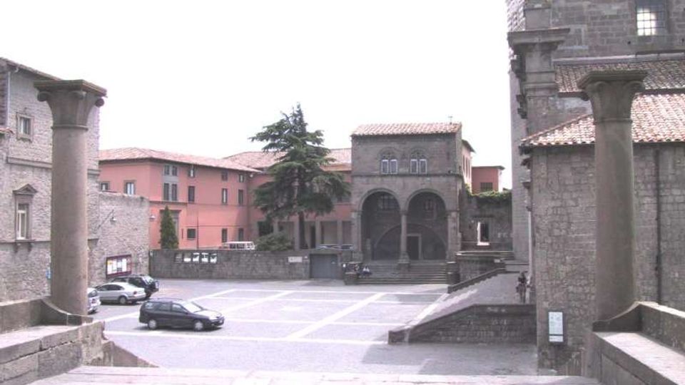 Piazza lastricata in un borgo storico, vista dall’alto di una scalinata in pietra con due colonne in primo piano. Attorno si vedono edifici in muratura con tetti in coppi, un porticato con archi, un grande albero centrale e alcune auto parcheggiate.