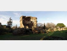 Rovine di una grande struttura circolare in pietra, con ampie parti crollate, situata in un’area erbosa. Intorno si vedono alberi e resti di murature più basse, sotto un cielo sereno.