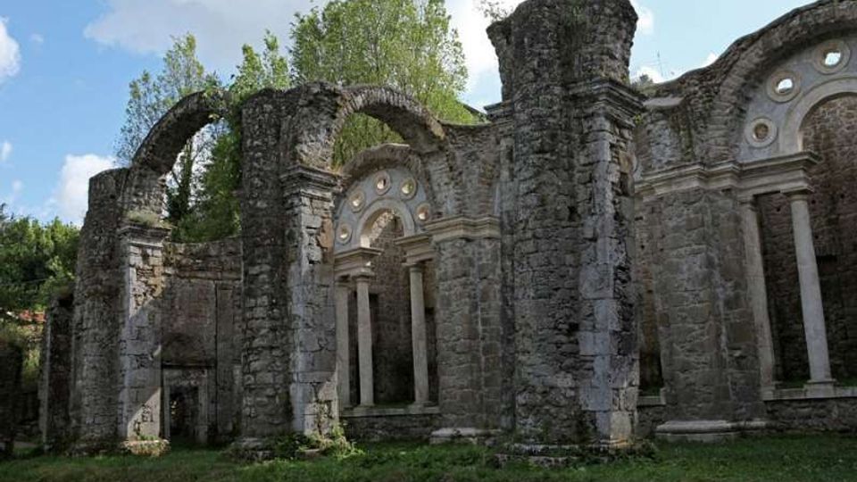 Rovine di un edificio in pietra, con grandi archi e colonne ancora in piedi e aperture ad arco con oculi circolari. L’area è circondata da vegetazione e alberi, con prato in primo piano e cielo visibile sullo sfondo.