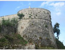 Torre circolare di una fortificazione in pietra con mura alte e spesse, vista dal basso su un pendio roccioso. Vegetazione spontanea cresce sulle pareti e intorno alla struttura, con cielo azzurro sullo sfondo.