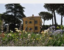 Villa in stile rurale dipinta di giallo con persiane verdi e tetto in coppi, circondata da un giardino fiorito in primo piano. Sullo sfondo si vedono alti pini e altri alberi sotto un cielo chiaro.