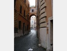 Vicolo urbano stretto con pavimentazione in sampietrini, fiancheggiato da alti edifici in muratura. In fondo, un arco in pietra collega i palazzi e incornicia una piazza con un edificio chiaro sullo sfondo.