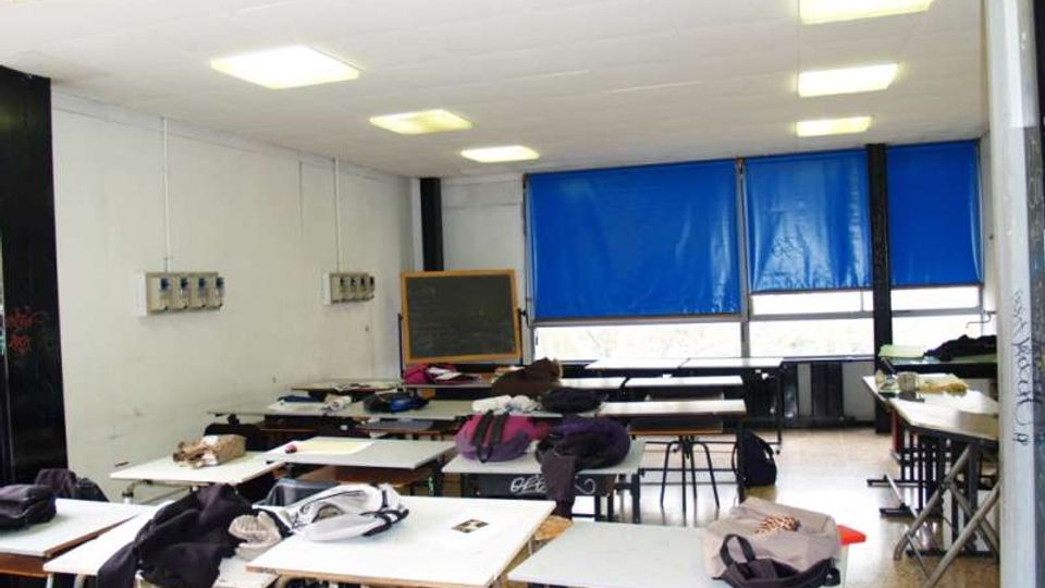 Aula scolastica con file di banchi e tavoli, soffitto con pannelli e luci rettangolari, e una lavagna sul fondo. Grandi finestre con tende blu lungo una parete illuminano lo spazio, con pareti bianche e pavimento in graniglia.