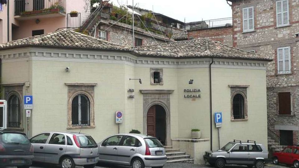 Edificio basso con facciata color crema e tetto in coppi, con ingresso ad arco e finestre ad arco con inferriate, con la scritta “Polizia Locale” sulla parete. Davanti si trova un’area di sosta con alcune auto parcheggiate e segnali blu di parcheggio, circondata da case in pietra su un lieve dislivello.