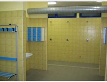 Spogliatoio con area docce, pareti rivestite di piastrelle gialle e pavimento grigio. Sono visibili un condotto di aerazione a soffitto, finestre alte con telaio blu, armadietti blu a parete e una panca metallica con appendiabiti.