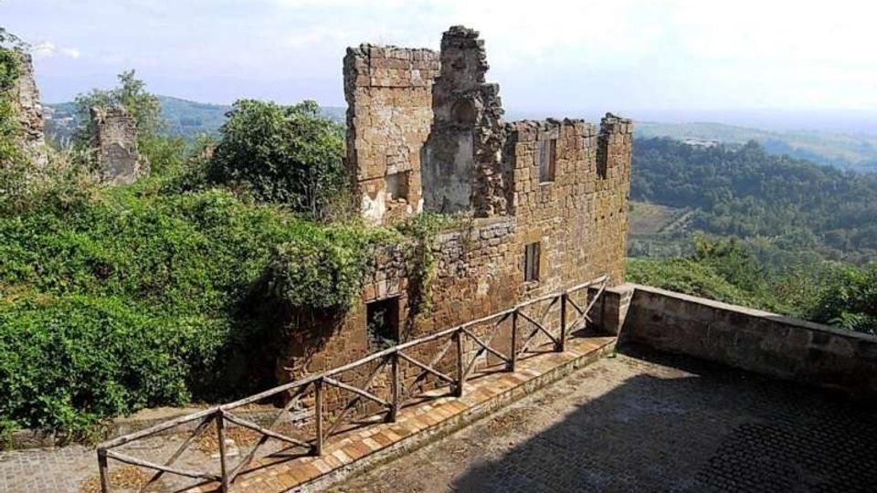 Ruderi di un edificio in pietra su un crinale, con muri parzialmente crollati e vegetazione che cresce intorno. In primo piano c’è una terrazza pavimentata con parapetto in legno e muretto, affacciata su colline boscose.