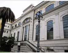 Facciata di un edificio storico con ampie finestre ad arco e cornici in pietra, con scalinata esterna e ingresso sotto un grande arco con scritta “SPQR”. In primo piano si vede un lampione ornamentale e vegetazione con palme, in un contesto urbano.