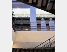 Interno di edificio industriale riconvertito, con passerelle metalliche e ringhiere su più livelli attorno a un grande vano centrale. Struttura in acciaio a vista, soffitto con travi scure e illuminazione diffusa dall’alto.